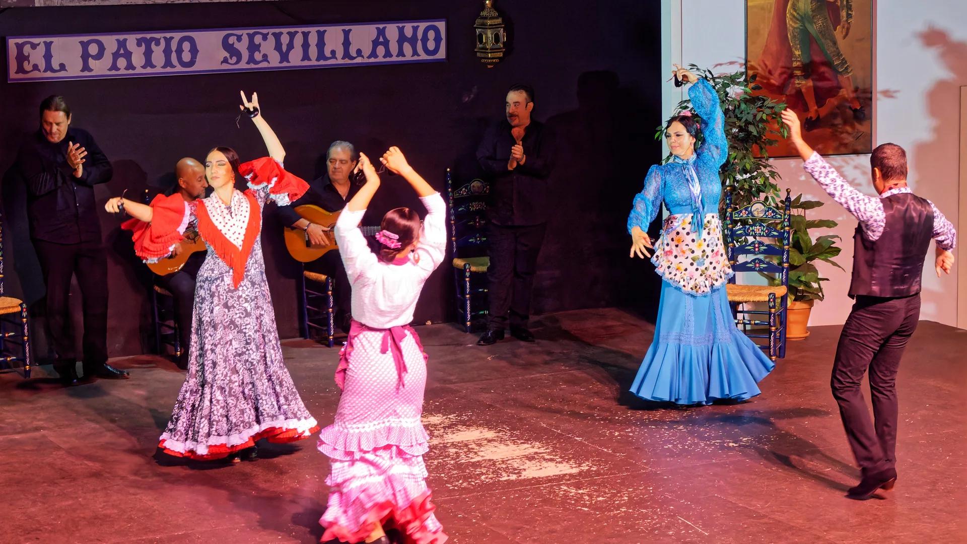 Flamenco dancers in red, pink and blue ruffled dresses raise castanets on a stage lit in red, with seated guitarists behind them and a blue tile sign reading El Patio Sevillano