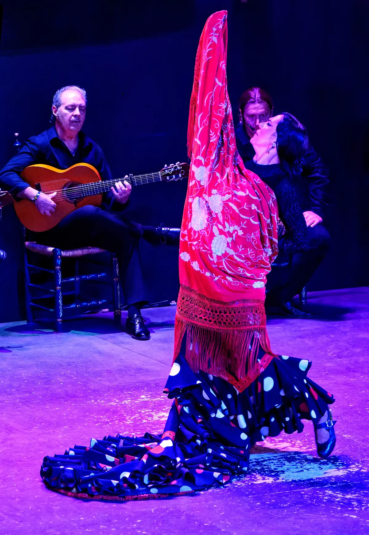 A dancer in a black polka-dot dress with a red shawl draped from her raised arms